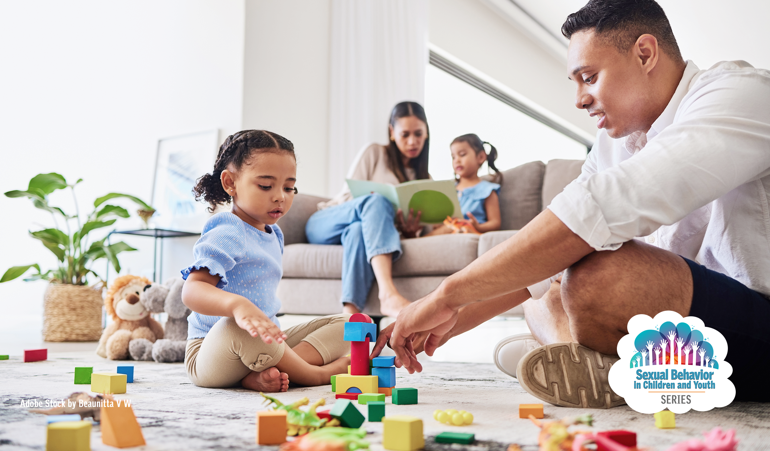Family, mother and girl on couch reading while father and other daughter sit on the ground learning and building toy blocks in front of the couch