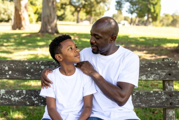 Image of a black father and ten year old son sitting on a bench; the father has his arm wrapped around the son's shoulders in a caring engaged way