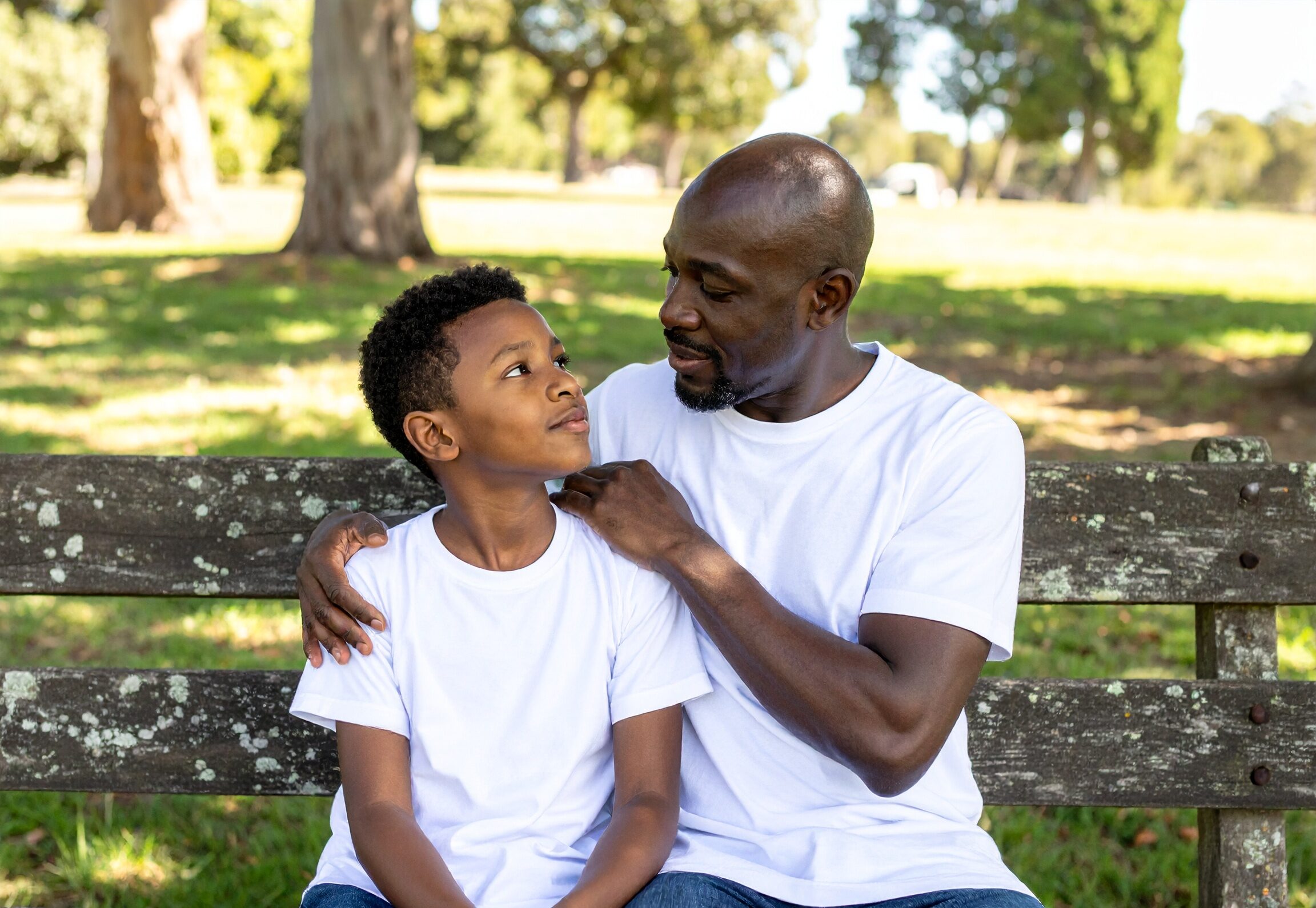 Image of a black father and ten year old son sitting on a bench; the father has his arm wrapped around the son's shoulders in a caring engaged way