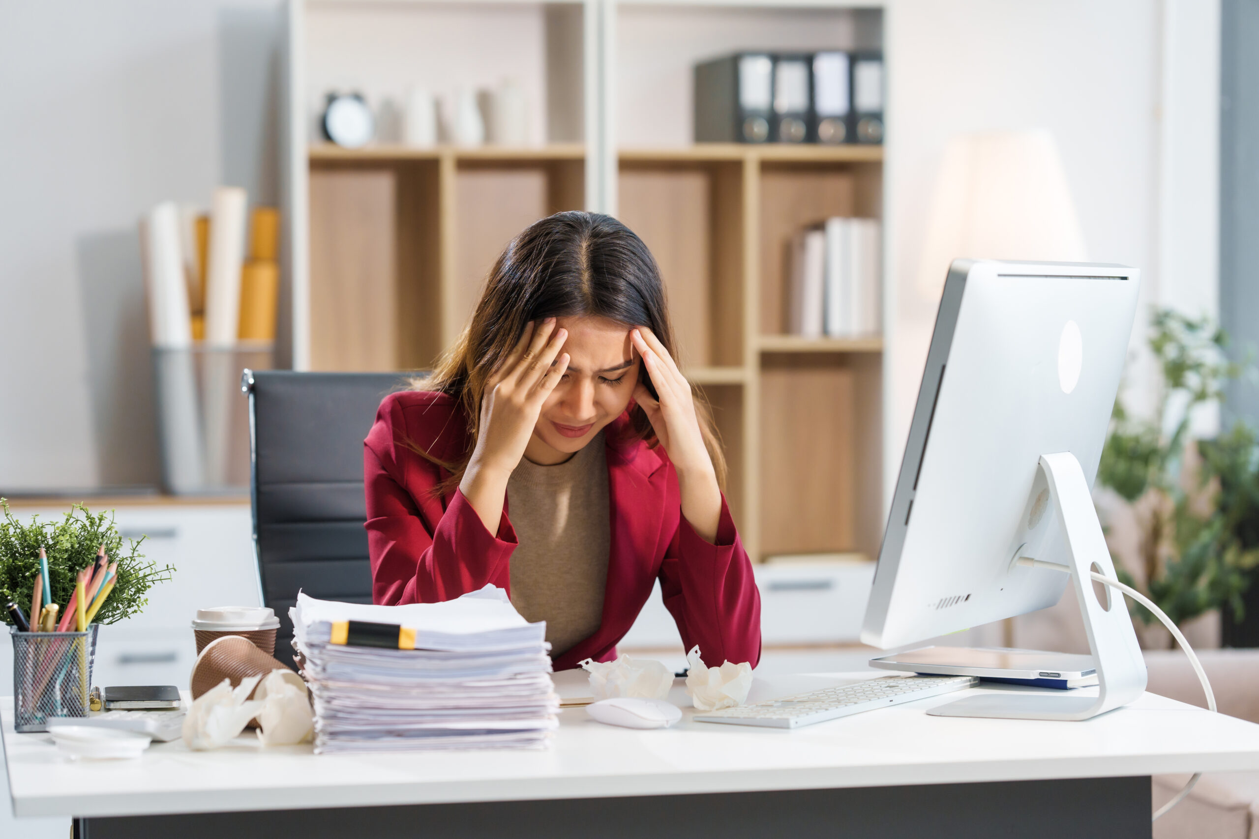 A professional woman sitting at a desk, visibly stressed and overwhelmed, resting her head in her hands on the table, showing signs of distress