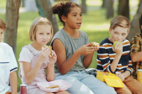 Diverse boys and girls of elementary school age sitting n bench outside eating a sandwich.