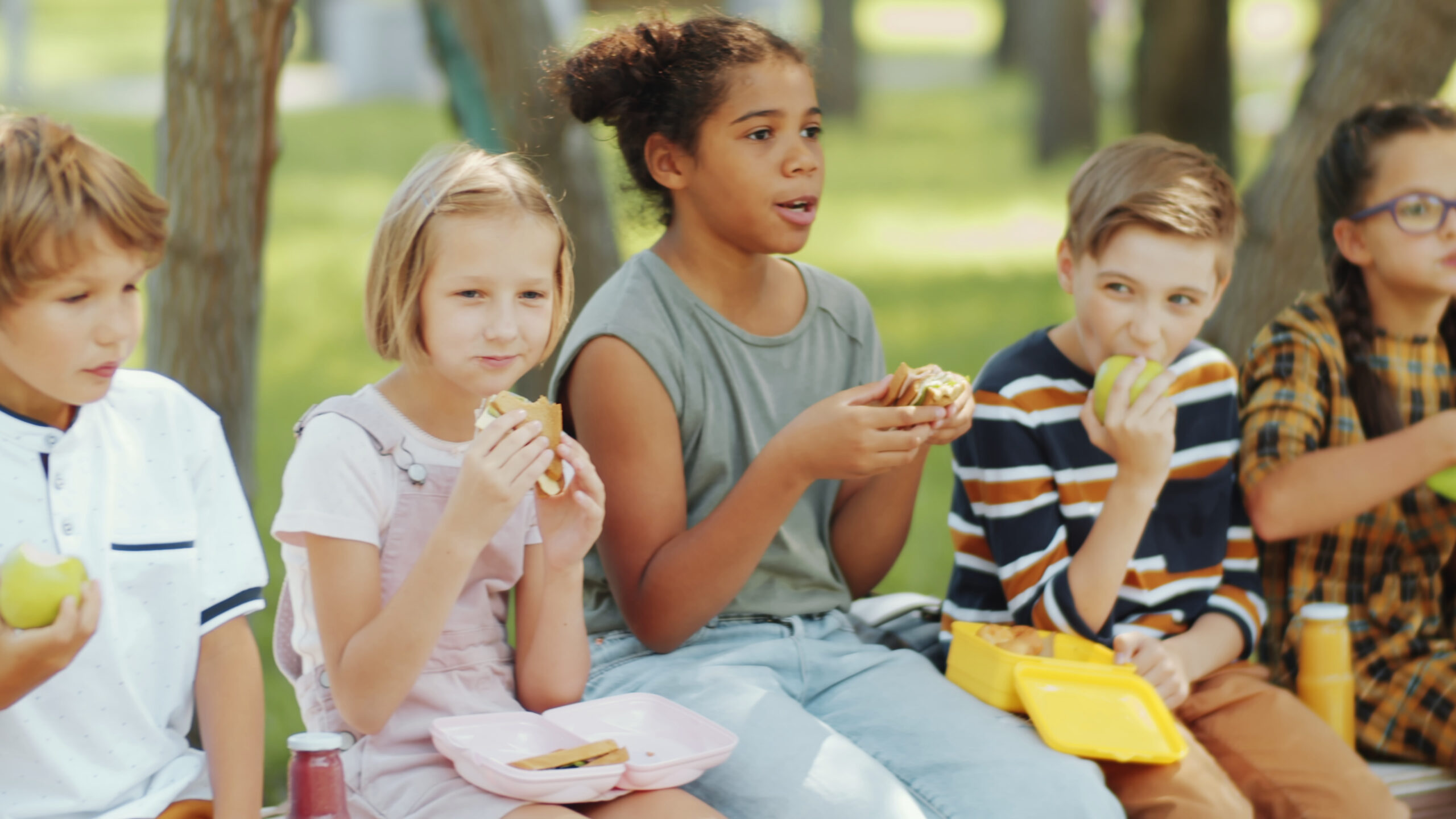 Diverse boys and girls of elementary school age sitting n bench outside eating a sandwich.