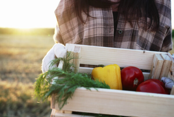 Woman holding a wooden box of fresh produce including carrots and sweet bell peppers fresh from the garden.