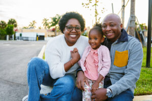 An African American couple and their three year old daughter sit on the curb in the parking lot of a public park in Florida at sunset and smile directly at the camera.