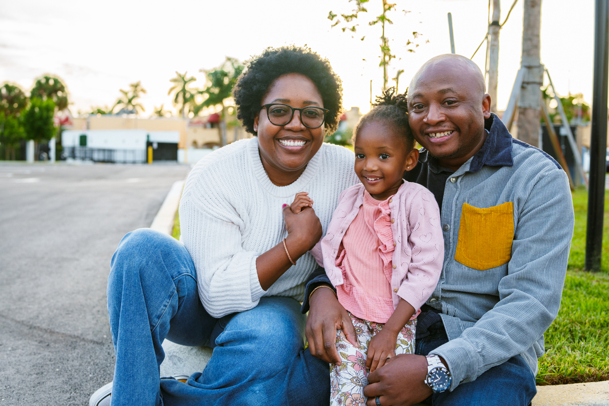 An African American couple and their three year old daughter sit on the curb in the parking lot of a public park in Florida at sunset and smile directly at the camera.