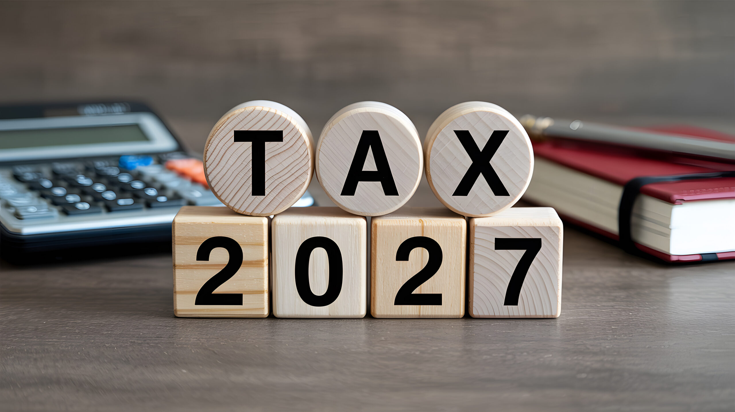 A stack of wooden blocks with tax 2027 written on them on a desk with calculator and book