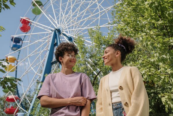 two teenagers standing in front of a ferris wheel, smiling and looking at each other